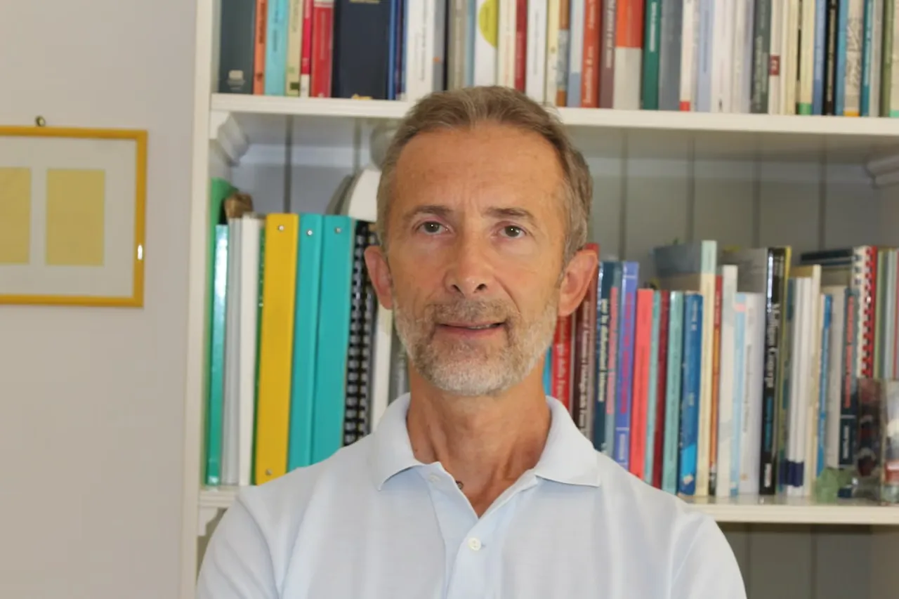 Uomo con barba grigia e maglietta azzurra davanti a una libreria piena di libri colorati.